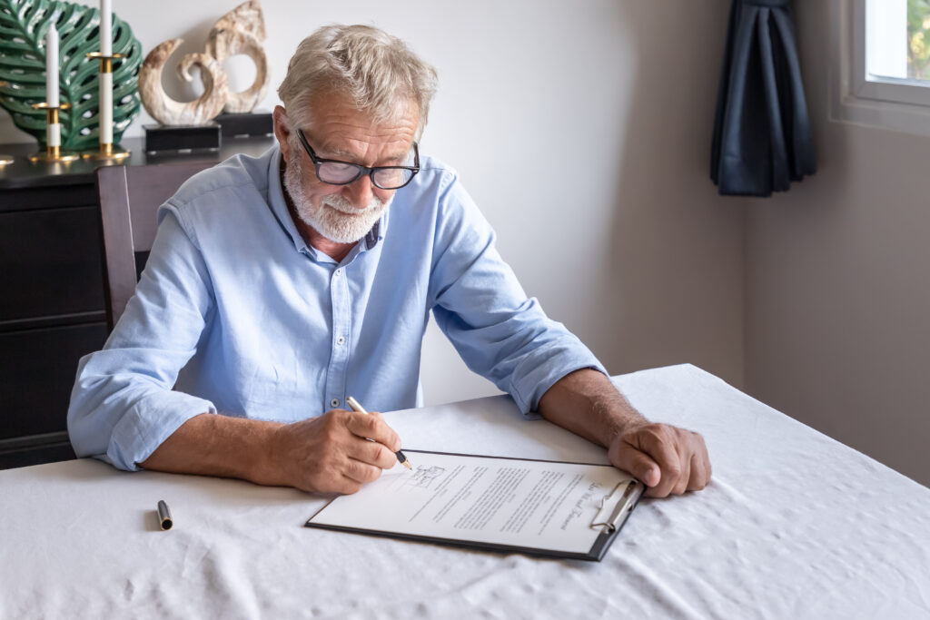 An elderly man sits at his dining room table and signs his Will.