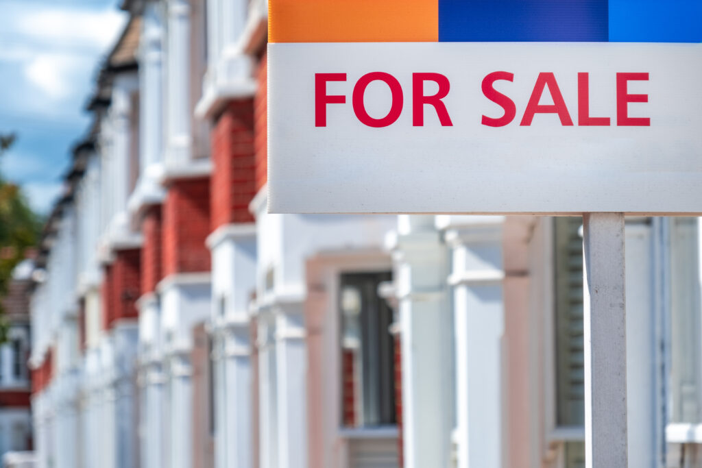 A closeup of a "for sale" sign outside of a row of terraced houses on a sunny day.