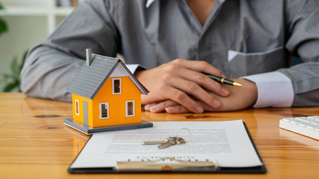 A model of a house and house keys placed upon a contract. A client prepares to sign the contract.