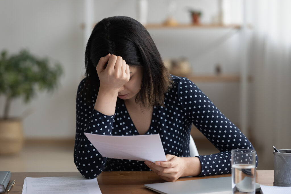 A woman appears frustrated and upset and holds her hand to her head as she reads a document.