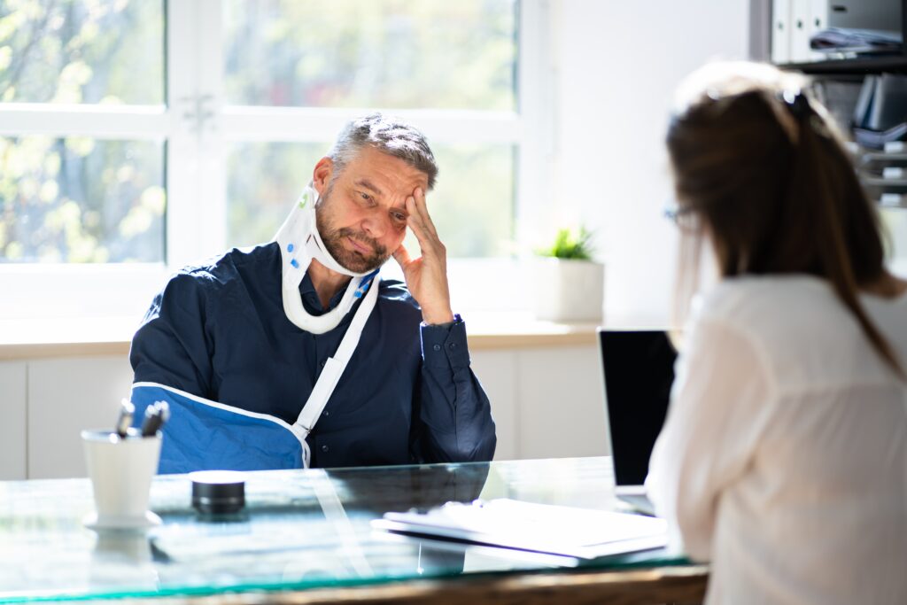 An injured man with a neck brace and arm sling consults with a female solicitor in her office.