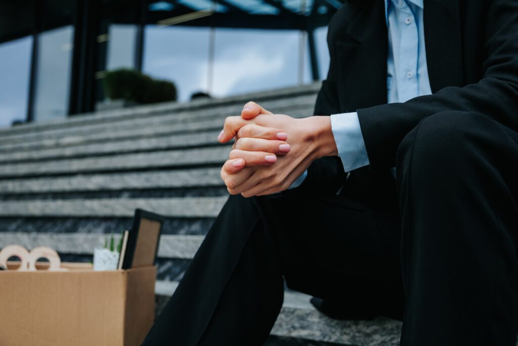 A businesswoman sorrowfully sits outside of an office with a box of her possessions after losing her job.