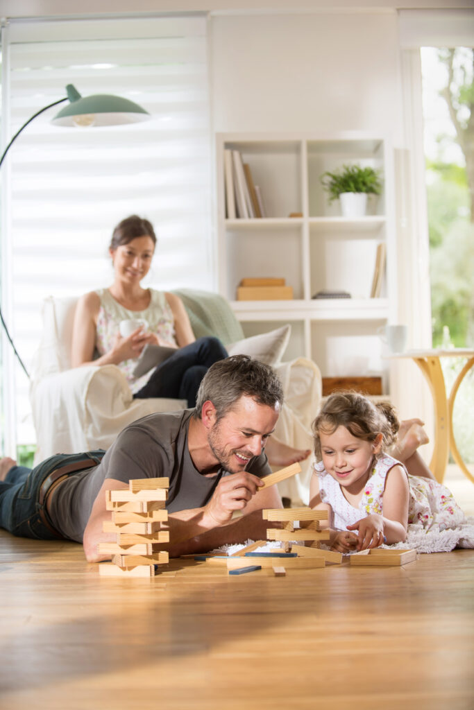 A cheerful young family relax in their living room. A father plays a game with his young daughter while his wife reads a book.