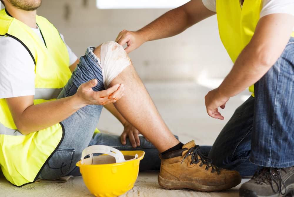 A construction worker wraps a bandage around his colleague's injured leg.